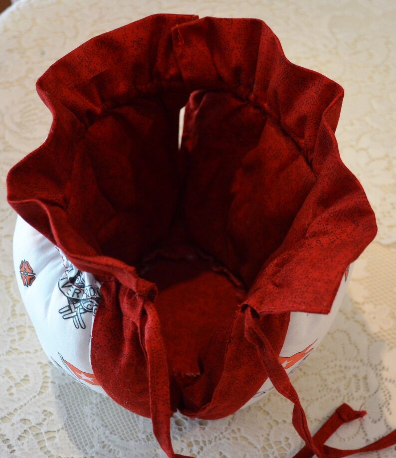 Puede incluir: Una bolsa de tela roja con cord&oacute;n de ajuste, forro blanco y base de tela blanca. La bolsa tiene un borde superior con volantes y un cierre de cord&oacute;n. La base de la bolsa tiene un patr&oacute;n de rayas blancas y rojas.