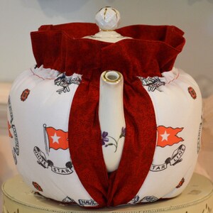 Puede incluir: Calienta-tetera blanca con una cubierta de tela roja y un dise&ntilde;o de estrella blanca. El calienta-tetera tiene el texto "White Star Line" y "NAVIC" en &eacute;l.