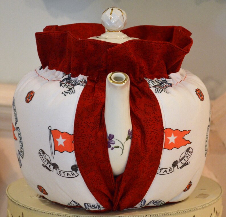 Puede incluir: Calienta-tetera blanca con una cubierta de tela roja y un dise&ntilde;o de estrella blanca. El calienta-tetera tiene el texto "White Star Line" y "NAVIC" en &eacute;l.