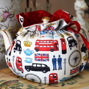 K&ouml;nnte beinhalten: Eine wei&szlig;e Teekannenhaube mit rotem Besatz und Kordelzug. Das Muster zeigt Londoner Ikonen wie rote Doppeldeckerbusse, schwarze Taxis und Union Jacks. Die Teekanne hat einen goldenen Knauf.