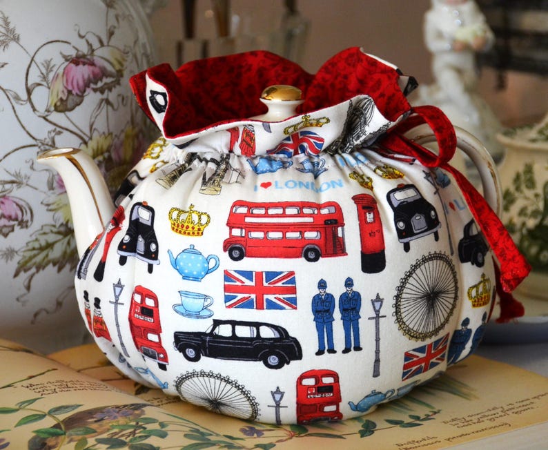 K&ouml;nnte beinhalten: Eine wei&szlig;e Teekannenhaube mit rotem Besatz und Kordelzug. Das Muster zeigt Londoner Ikonen wie rote Doppeldeckerbusse, schwarze Taxis und Union Jacks. Die Teekanne hat einen goldenen Knauf.