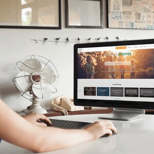 以下が含まれることがあります： 女性が机に座ってコンピューターの画面の前にいます。画面には「探検して発見」というテキストと検索バーが表示されています。ウェブサイトには、野原を歩く家族の写真と「ブログを閲覧する」というタイトルのセクションも表示されています。