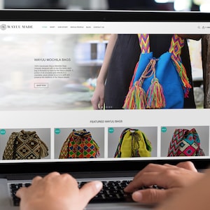 May include: A laptop computer displaying a website for Wayuu mochila bags. The website features a blue bag with colorful tassels and a section showcasing featured bags in various colors and patterns. The text on the website reads "Wayuu Mochila Bags" and "Featured Wayuu Bags".