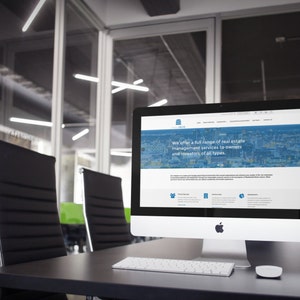 May include: A modern office setting with a black desk and two black leather chairs. A white iMac computer is on the desk with a website open on the screen. The website is for a real estate management company and features a cityscape background with the text "We offer a full range of real estate management services to owners and investors of all types."