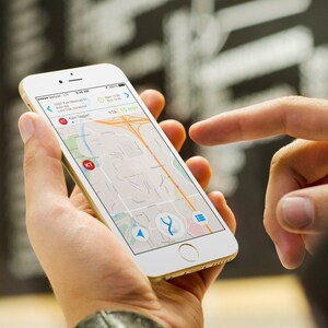以下が含まれることがあります： 人がスマートフォンを使って地図アプリでナビゲートしています。画面には、青いルートと「KT」というラベルの付いた赤いマーカーが表示された地図が表示されています。アプリは、到着予定時刻を「15分」と表示しています。