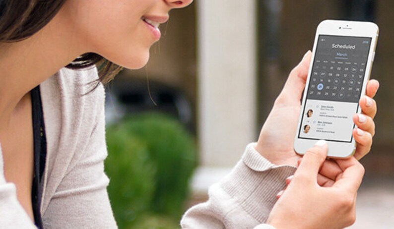 以下が含まれることがあります： スマートフォンを使ってカレンダーアプリを見ている人がいます。アプリは3月のカレンダーを表示しており、27日に青い点が付いています。アプリは、名前と時間付きのスケジュールされた予定のリストも表示しています。