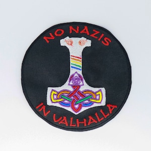 Könnte beinhalten: Runder, gestickter Aufnäher mit schwarzem Hintergrund. Das Design zeigt einen weißen Hammer mit Regenbogenstreifen und einem violetten Knoten. Die Worte "NO NAZIS" und "IN VALHALLA" sind rot gestickt.