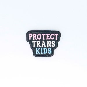 May include: Black fabric patch with the text "PROTECT TRANS KIDS" in pink, white, and blue lettering.
