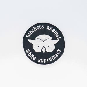 Puede incluir: Un parche redondo negro con texto bordado blanco y un diseño de búho blanco. El texto "teachers against white supremacy" está dispuesto en círculo alrededor de la ilustración del búho. El parche está centrado sobre un fondo blanco.