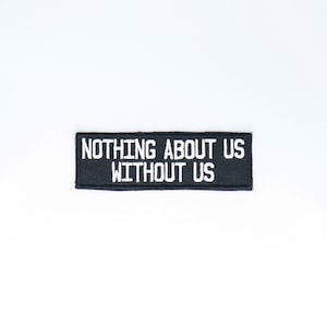 May include: Black embroidered patch with white text that reads "Nothing About Us Without Us".