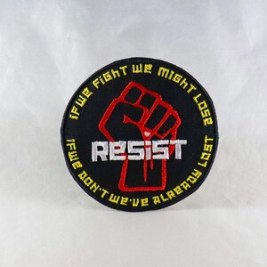 Op de afbeelding: Zwarte en gele geborduurde patch met een rode vuist en de tekst "If we fight we might lose if we don't we've already lost" en "Resist" in wit.
