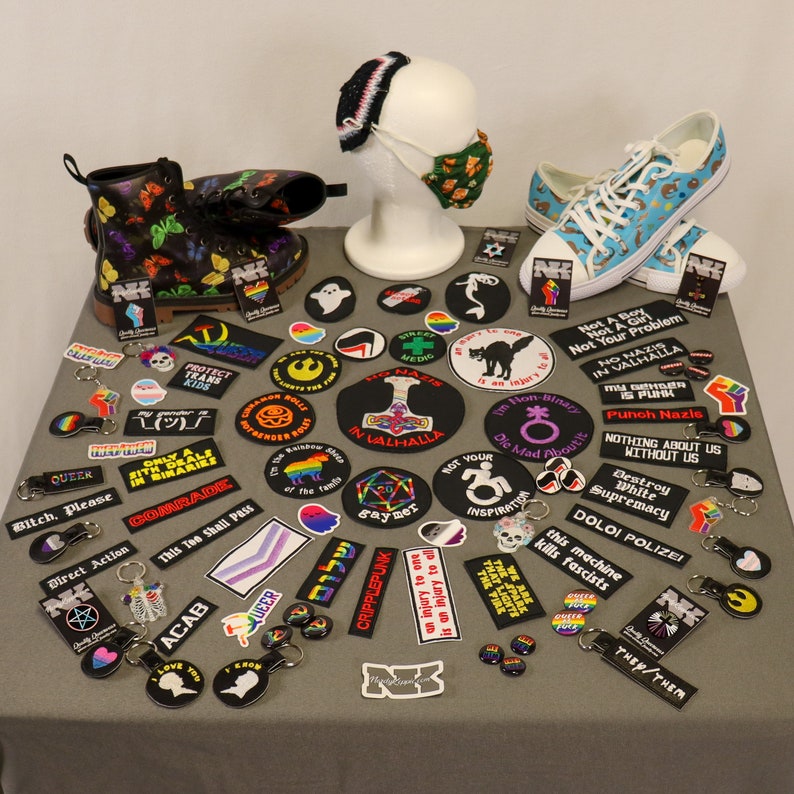 Pu&ograve; includere: Una collezione di toppe ricamate con vari slogan e disegni, tra cui "This Too Shall Pass", "Punch Nazis", "Destroy White Supremacy" e "Only a Sith Deals in Binaries". Le toppe sono disposte su una superficie bianca con altri accessori, come portachiavi e un paio di scarpe da ginnastica bianche e nere.