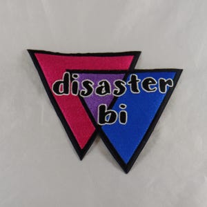 May include: Embroidered patch featuring two overlapping triangles in pink and blue, with a purple center. The patch reads "disaster bi" in black letters with a white outline. The patch has a black border.
