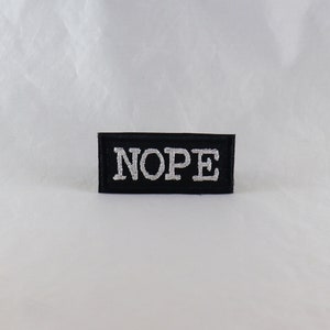 Puede incluir: Un parche rectangular negro con la palabra "NOPE" bordada en hilo plateado. El parche es una declaración simple y audaz, perfecta para agregar a la ropa o accesorios.