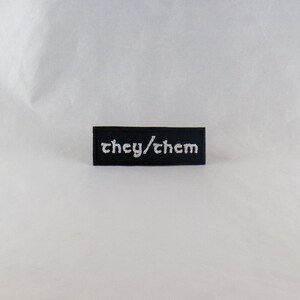 Könnte beinhalten: Schwarzer gestickter Aufnäher mit weißem Text, der "they/them" lautet.