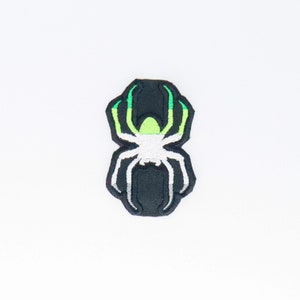 Puede incluir: Un parche bordado de araña en negro y blanco con detalles en verde y verde claro. La araña está en una forma de telaraña.