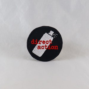 May include: A black and white embroidered patch with a spray paint can and the text "direct action" in red.