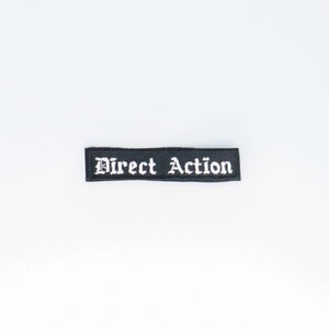 Può includere: Toppa in tessuto nero con testo ricamato bianco che recita "Direct Action".