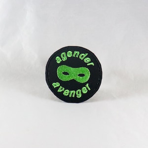 Może przedstawiać: Czarna i zielona naszywka haftowana z tekstem "agender avenger" i zieloną maską na oczy.