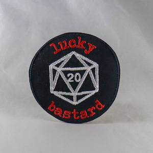May include: Black circular patch with red embroidered text reading "lucky bastard". A white embroidered 20-sided die is in the center, with the number 20 in the middle. The patch is likely for a gamer or role-playing enthusiast.