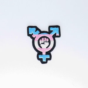 May include: A blue, pink, and white transgender pride flag with a raised fist in the center, embroidered on a black patch.