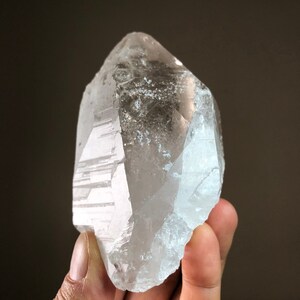 May include: A large, clear quartz crystal with a pointed tip and a rough, natural surface. The crystal is held in a hand, showcasing its size and clarity.