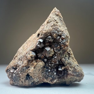 May include: A rough, brown rock with a cluster of dark brown, faceted crystals. The crystals are arranged in a random pattern and appear to be embedded in the rock.