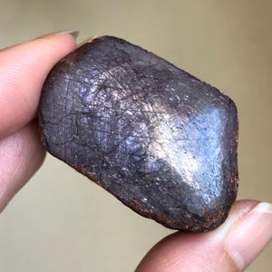 May include: A polished, rectangular-shaped gemstone with a deep, dark purple and brownish-red hue. The surface displays a complex network of fine, intersecting linear inclusions and a subtle, reflective sheen across its textured face.