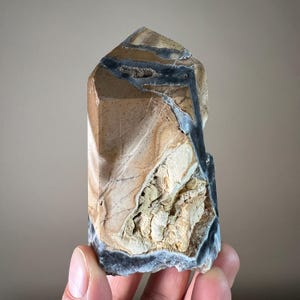 May include: A polished stone obelisk with a natural, earthy color palette. The stone features shades of tan, brown, and black, with intricate veining and patterns. The top is angled, and the base is rough-hewn. A unique decorative item.