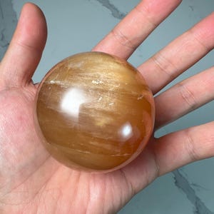 May include: A polished, spherical gemstone in shades of amber and gold. The orb has a smooth, reflective surface with visible internal patterns. The gemstone is held in an open hand, showcasing its size and color.