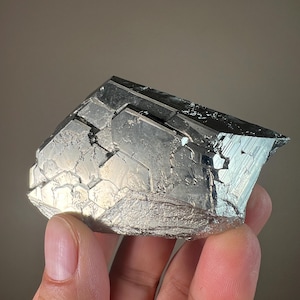 May include: A metallic, silver-colored mineral specimen with a geometric, crystalline structure. The surface reflects light, showcasing a complex pattern of facets and angles. The mineral appears to be held by a hand.