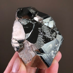 May include: A large, black, faceted hematite crystal with a metallic sheen. The crystal has a rough, natural texture and is composed of multiple facets.