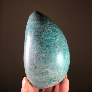 May include: A polished, teardrop-shaped stone in teal and blue-green hues. The smooth, glossy surface displays natural veining and subtle colour variations. The object is held in a hand against a dark brown background.