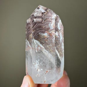 May include: A clear quartz crystal with a pointed top. The crystal has a layered appearance with brown and reddish-brown inclusions. The lower portion of the crystal is clear, while the upper section has a textured, opaque look. The crystal is held by a hand.
