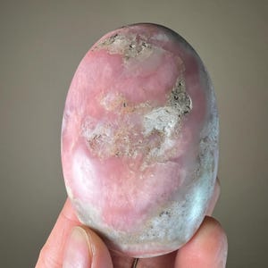 May include: A polished, oval-shaped pink rhodonite stone with areas of white and gray. The stone has a smooth surface and a natural, organic appearance. The pink color varies in intensity throughout the stone.