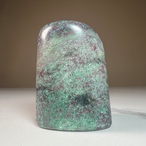 May include: A smooth, polished green and purple gemstone with a unique, swirling pattern. The stone is shaped like a rounded, asymmetrical pyramid.