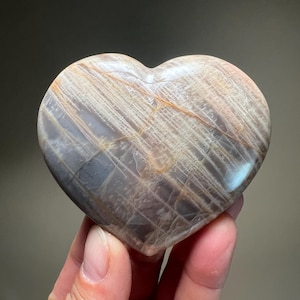 May include: A heart-shaped stone with a polished surface. The stone features a mix of colors, including gray, beige, and hints of orange. The surface has a pattern of parallel lines, creating a textured appearance. The stone is held in a hand.