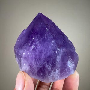 May include: A large, raw amethyst crystal with a deep purple hue. The crystal has a pointed, faceted top and a rough, textured surface. The gemstone is held in a hand, showcasing its natural beauty and crystalline structure.