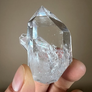 May include: A clear quartz crystal with a pointed tip and a rough, textured surface. The crystal is held in a hand and appears to be transparent, with light refracting through it. The crystal is a natural mineral specimen.