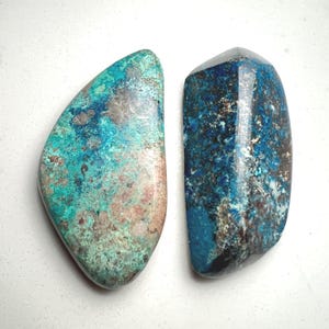 May include: Two polished gemstones with unique patterns. One is a mix of teal, blue, and brown, while the other features deep blue and black hues. The stones have irregular shapes and a glossy finish, perfect for jewelry or display.
