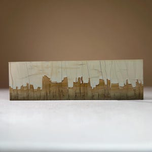 May include: A rectangular wooden block with a skyline design. The top section is a light beige, and the bottom has a brown, textured appearance. The skyline is cut out of the wood, creating a distinctive visual effect. The block is approximately 25 cm long.