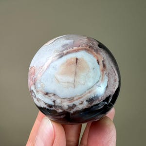 May include: A polished, spherical stone with a unique pattern. The orb features a mix of colors, including white, brown, black, and hints of pink. The stone's surface has a smooth, glossy finish, and the overall shape is perfectly round.