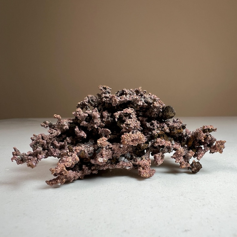 Pode incluir: Um esp&eacute;cime de cobre natural com uma superf&iacute;cie &aacute;spera e texturizada e uma cor castanho-avermelhada. A forma&ccedil;&atilde;o mineral tem uma estrutura complexa e ramificada, e est&aacute; sobre uma superf&iacute;cie branca. O fundo &eacute; de cor bege neutra.