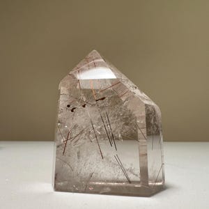 May include: A clear quartz crystal tower with reddish-brown and black inclusions. The crystal has a polished, faceted shape with a flat top and a rectangular base. The inclusions appear as thin lines and streaks within the transparent quartz.