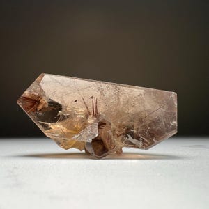 May include: A clear, pale brown crystal with reddish-brown inclusions and a small, dark brown crystal base. The crystal has a geometric shape with sharp edges and a polished look. The background is a gradient of dark brown to white.