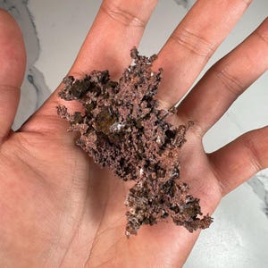 Pode incluir: Um peda&ccedil;o de min&eacute;rio de cobre bruto, segurado em uma m&atilde;o. O cobre tem uma superf&iacute;cie &aacute;spera e texturizada com uma cor marrom avermelhada. Algumas &aacute;reas mostram um tom mais escuro, quase preto, com pequenas manchas brancas.