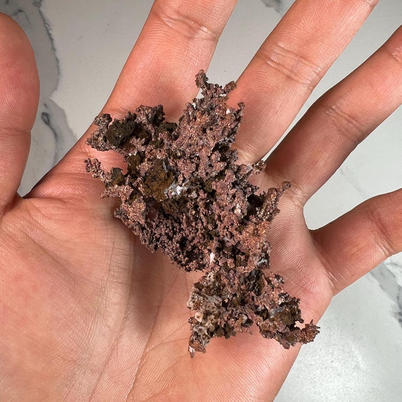 Pode incluir: Um peda&ccedil;o de min&eacute;rio de cobre bruto, segurado em uma m&atilde;o. O cobre tem uma superf&iacute;cie &aacute;spera e texturizada com uma cor marrom avermelhada. Algumas &aacute;reas mostram um tom mais escuro, quase preto, com pequenas manchas brancas.