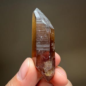 May include: A faceted, brown crystal with a pointed tip. The crystal has a translucent quality, with visible internal structures and a gradient of color from light amber to dark brown. The crystal is held in a hand.