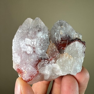 May include: A cluster of clear quartz crystals with red and white inclusions. The crystals have a rough, textured surface and are held in a hand. The overall appearance is natural and geological.