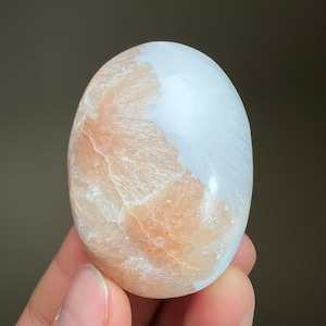 May include: Oval-shaped polished stone with a smooth, glossy surface. The stone features a blend of peach and white colors, with a unique, swirling pattern. The peach-colored section has a textured, almost crystalline appearance.